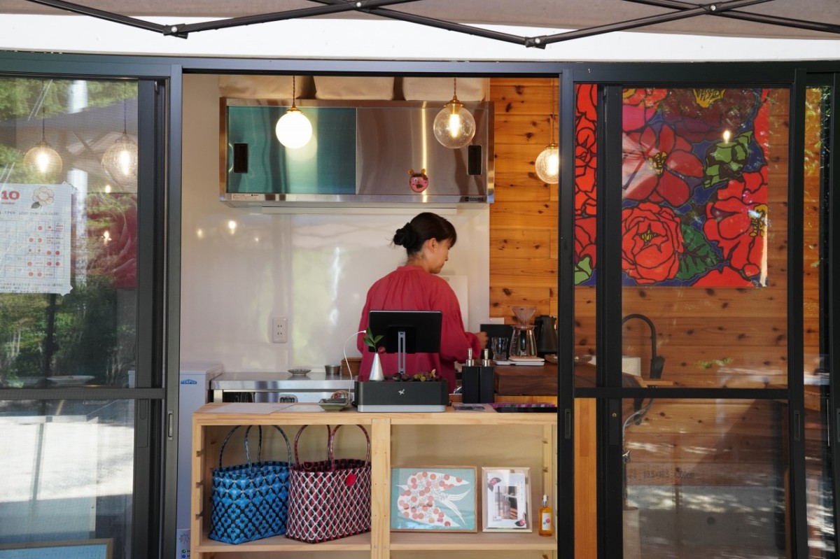 椿の絵が飾られたキッチンで、赤い服を着て準備する椿の森カフェの店主